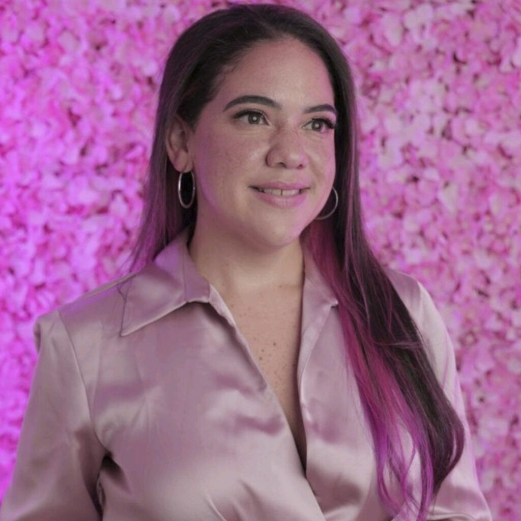 Especialista en uñas Esteffanie Díaz, sentada elegantemente con un fondo rosado, lista para enseñar cursos de diseño de uñas en línea y presenciales